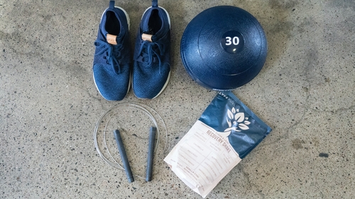On grey concrete sits a pair of blue running shoes with white details with a white exercise ball with the number 30 on the top in white sitting next to them. Also on the floor is a jump rope with blue/grey handles and a blue packet of protein powder.