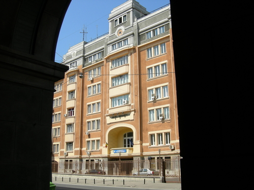 The Bucharest Police Headquarters is visible from across the street, framed on either side by arched shadows. The building is seven or eight storeys tall, and the outside walls are a light pinkish brown.