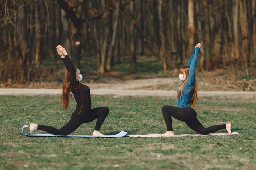 two women in a field doing warriors pose (yoga) They are kneeling on the ground with their arms in the air, facing each other. The woman on the left id wearing all black and has long brown hair. The woman on the right is wearing light blue and black and has blond hair. Both are wearing disposable masks. There is a forest visible in the background.