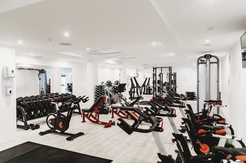 A gym is shown with light wooden floors and white walls and ceiling. In the gym is numerous workout equipment and machines such as spin bikes. All the machines are black with orange details.