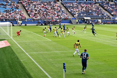 A football game is happening in a stadium full of people on the stands. One team is wearing green/yellow and white uniforms while the other team is wearing blue uniforms. A blue team member is in the middle of kicking the football towards the other teams goal. The goalie is wearing red is looks ready to stop the goal.