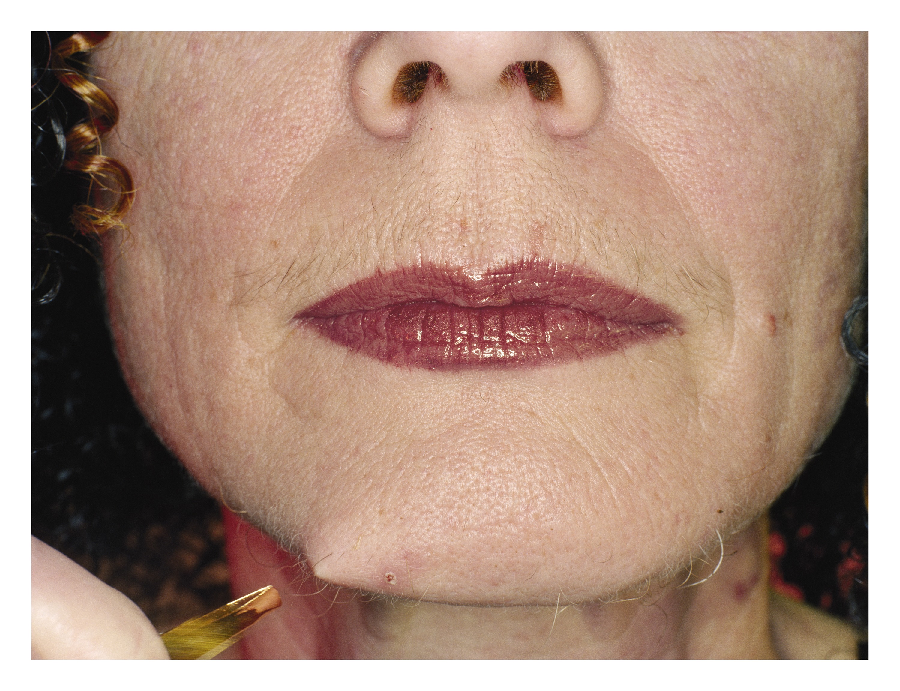 Colour photo of feminine appearing person plucking a hair on their chin. They are wearing lipstick and in the top left corner their hair can be seen curled. 