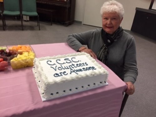 Colour photo of a volunteer at the Chalmers Community Services Centre sitting at a table covered by a pink tablecloth with a fruit tray on it and a white cake with "CCSC volunteers are awesome" written on it in icing.