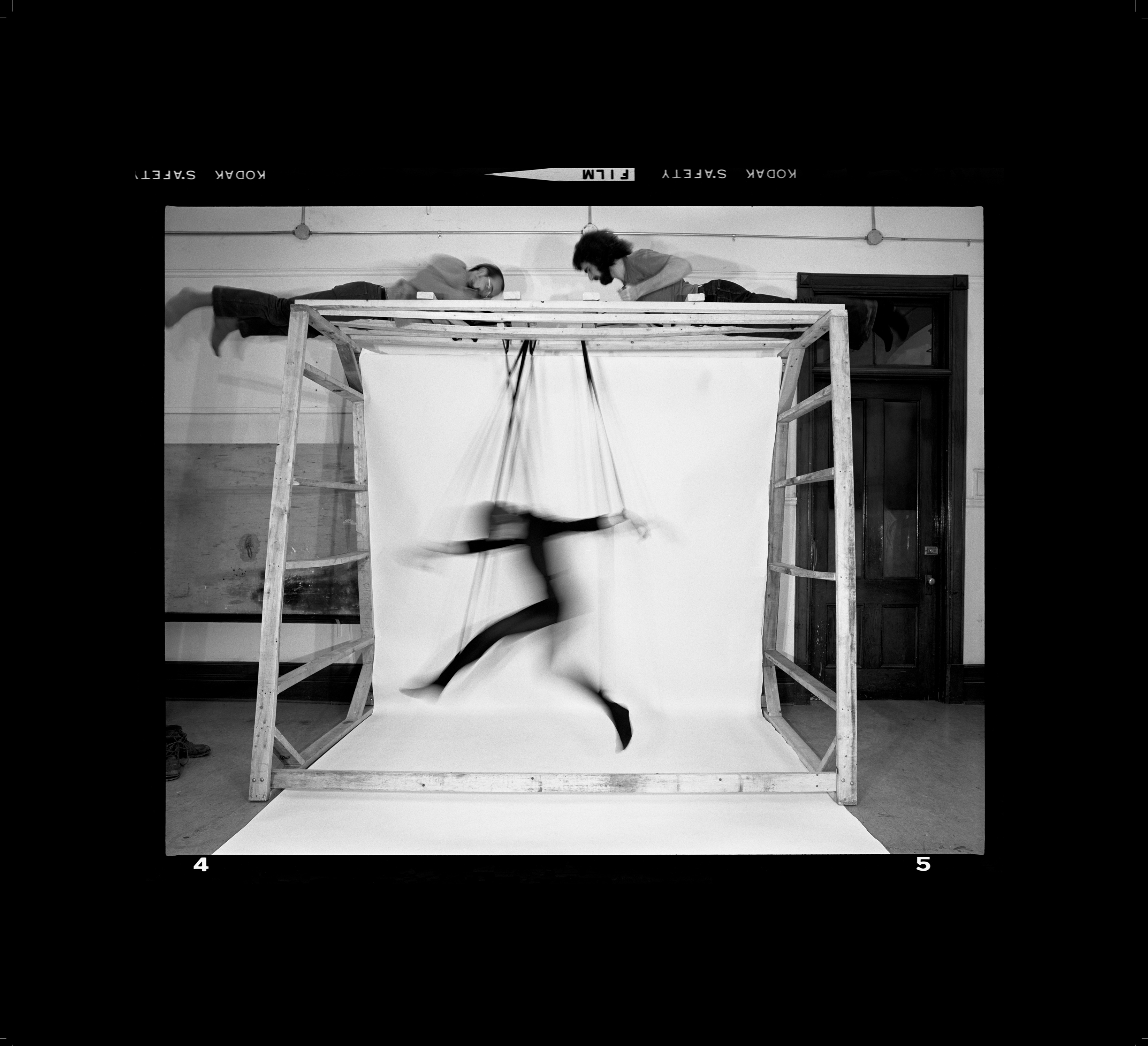 Black and white photo of Choreographed Puppet #4.5 by Suzy Lake. This artwork depicts a body form suspended by wires, imitating that of a puppet. The background appears to be some sort fo room with painted bricks similar to that of the inside of a school.