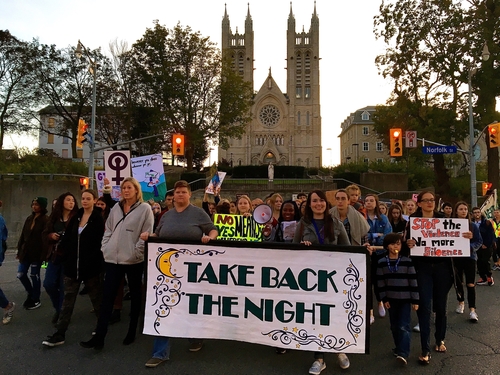 Colour photo of Take Back the Night Marchin Guelph on the street in front of the Church of Our Lady Basilica. There is a large crowd that is carrying a banner that says "TAKE BACK THE NIGHT" with a large yellow moon in the top left.