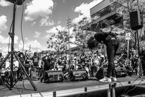A black and white image of Audny-Cashae Stewart bowing in front of a crowd with her arms crossed behind her back, holding a microphone. The crowd of people wear masks and hold signs. The crowd is active and engaged, many raising their hands or taking pictures with phones or cameras.