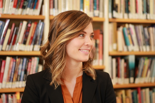 Dr. Amy Ellard-Gray standing in front of a row of bookshelves, smiling and looking off to the right. Dr. Ellard-Gray is wearing an orange blouse with a black blazer.