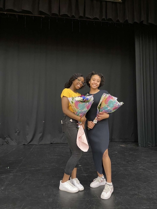 Audny-Cashae Stewart (right) and friend Carliene Christian (left) pose together, each holding a bouquet of brightly-wrapped flowers. They both smile at the camera and stand on a black auditorium stage in front of black curtains.