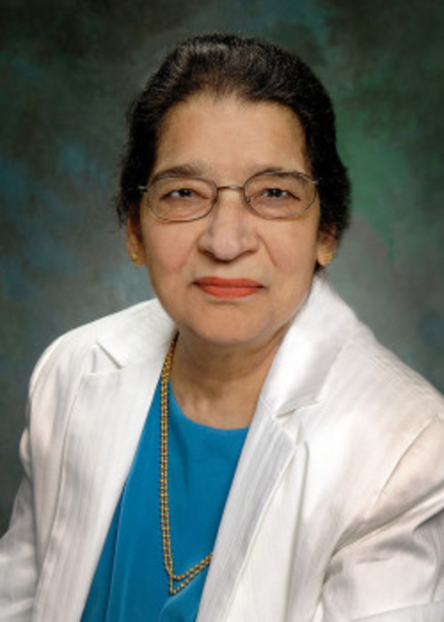 Professional photograph of Dr. Parvathi Basrur. She is facing the camera in front of a standard photography background. She is wearing a white suit jacket, with a turquoise shirt underneath, and a gold necklace is around her neck. 