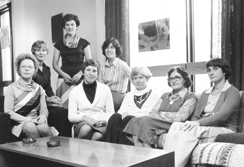 Black and white photo of the CFUW Guelph Executive team, 1970. In this photograph are shown Fran Struthers (recording secretary), Janet McLean (president), Cathy Burns (past president), Marina Zitnak (social convenor), and Norma Neudoerffer (publicity). In the back row are Betty Dewhirst (program convenor), Joan Jenkinson (corresponding secretary), and Judy McVittie (vice president). Photo appears courtesy of the Guelph Public Library archives, Executive members, (F4-04-0-4-2) 1977. From left to right: there is a person sitting with her hands in her lap wearing a sweater with four lines diagonally across it, she is wearing glasses and their hair is curled. The woman to the right of them is wearing a dark colour suit and her hair is cut short, the woman to the right of her is wearing a pantsuit, above her near the middle of the group, there is a woman wearing a dark colored shirt with a scarf and earrings. The woman to the right of her is wearing a striped shirt. The woman in front of her to the right is wearing a scarf and pantsuit. The woman to her right is wearing a button up shirt underneath a vest and a necklace. The woman to the right of her is wearing a pantsuit. They are sitting behind a table in front of a large window with the curtains drawn and light coming in.