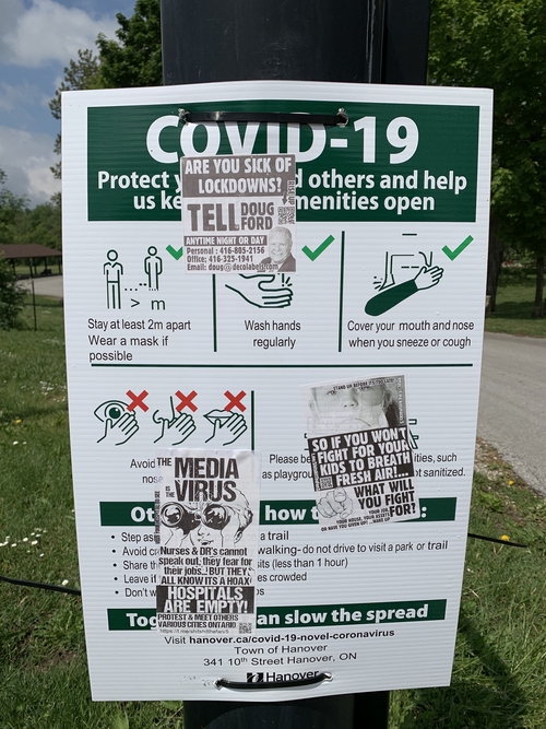 A white and green COVID-19 safety regulations sign, approximately 2 feet tall, attached to a gate post vandalized by 3 different COVID-19 disbelievers propaganda stickers at a community park and children's playground. The first sticker on the top left corner of the sign reads "Are you sick of lockdowns? Tell Doug Ford anytime night or day" and then lists the Ontario Premier's personal and business phone numbers, his email address, as well as a picture of Doug Ford. The second sticker at the middle right side of the sign reads "So if you won't fight for your kids to breath fresh air!...what will you fight for? Your job, your house, your assets or have you given up? ...wake up" with an image of a finger pointed at the reader. The third sticker towards the bottom lefthand side of the sign reads "The media is the virus. Nurses and DR's [doctors] cannot speak out, they fear for their jobs...but they all know it's a hoax. Hospitals are empty! Visit your hospital to see. Protest and meet others; various cities Ontario" with an image of a man holding binoculars to his eyes. All 3 stickers are in black and white colouring with QR codes to scan with a cell phone app to "Rise Up".