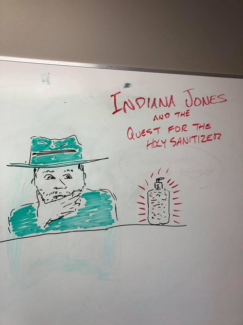 A sketch on a whiteboard of a man looking at a sparkling bottle of hand sanitizer. Text above the image reads "Indiana Jones and the Quest for the Holy Sanitizer." The sketch is a satire of the opening scene of "Indiana Jones and the Raiders of the Lost Arc" where Jones is scratching his chin while looking at a golden idol.