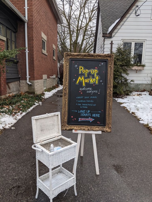 A photograph of a chalkboard sign that reads: "Pop-Up Market: welcome everyone! Wear your mask, sanitize your hands, one household at a time." Beside the sign is a basket containing a bottle of hand sanitizer and same COVID-19 face masks. The sign is located on the driveway of the home in Kitchener, Ontario, which hosted a socially-distanced art market before the holidays.