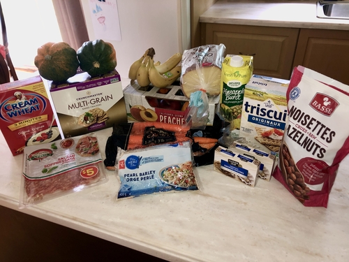 Grocery haul on a white countertop with tan cabinets and a white refrigerator in the background. From left to right, the food consists of: one box of Cream of Wheat, a package of Hot Salami, two small squash, a box of Multi-Grain Baked Crackers, four bananas, a tray of California Grown Peaches, a bag of big carrots, a bag of Pearl Barley, a bag of Artesian Bread, Pineapple Burst juice carton, box of Original Triscuits, two packages of Smoked Oysters, bag of Raw Hazelnuts.