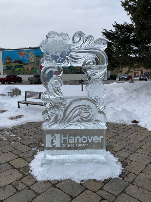 Taken in Hanover town square, the town mural in the background. An ice sculpture (approximately 6 feet tall) for the town's Family Day celebrations. The sculpture has a solid ice base, roughly 2 feet tall, that has the words "Hanover, Ontario, Canada" with the town's logo, with "#INTHISTOGETHERHANOVER" beneath it, hidden slightly by the snow the sculpture is resting on. The top of the sculpture resembles a picture frame made of elegant swirls and snowflakes, a particularly large snowflake in the large top left corner of the sculpture. There is a square hole cut in the center of the swirled/snowflake frame so citizens can take photos with the sculpture.