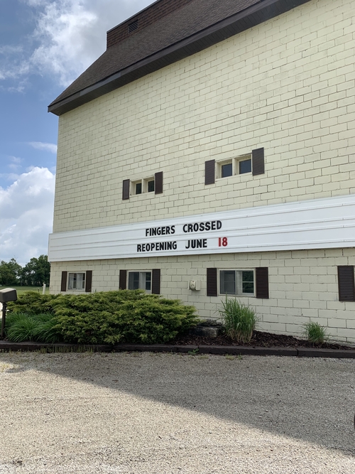 In front of the local Drive-In movie theatre, a tall white brick building. The advertising board where movie titles are displayed reads "Fingers Crossed, Reopening June 18th" in response to the hope that Ontario will begin phase 1 of reopening in the coming weeks.